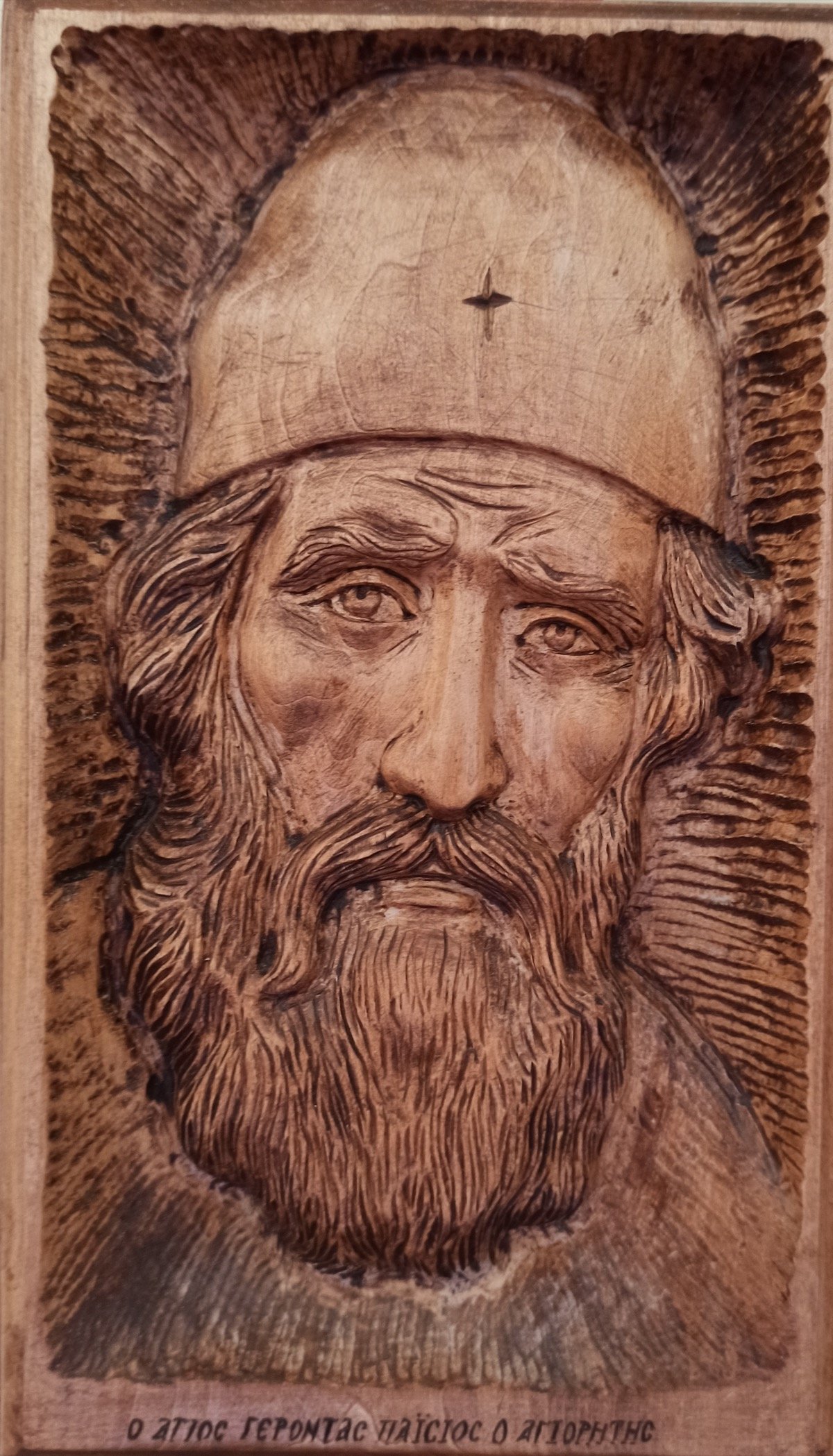 Wood-carved portrait of St. Paisios the Athonite, showing his distinctive monastic cap and gentle, weathered face, with Greek inscription