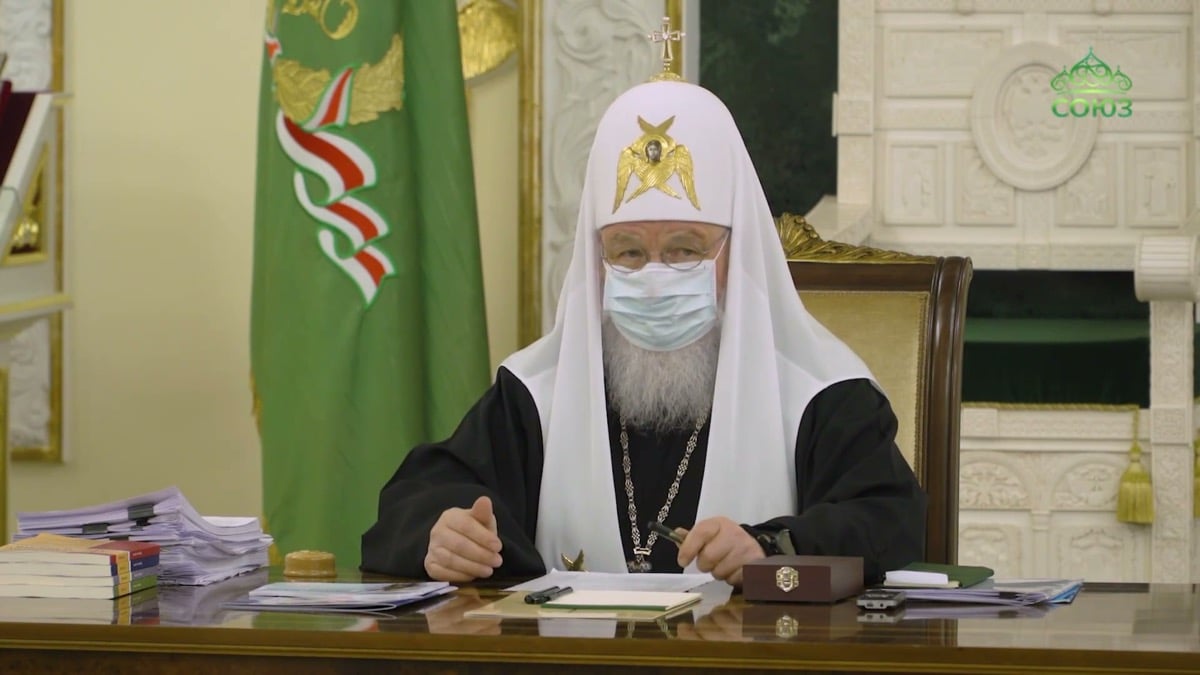 Patriarch Kirill wearing a surgical face mask and white patriarchal klobuk, seated at his desk at the Holy Synod meeting in Moscow, April 2021