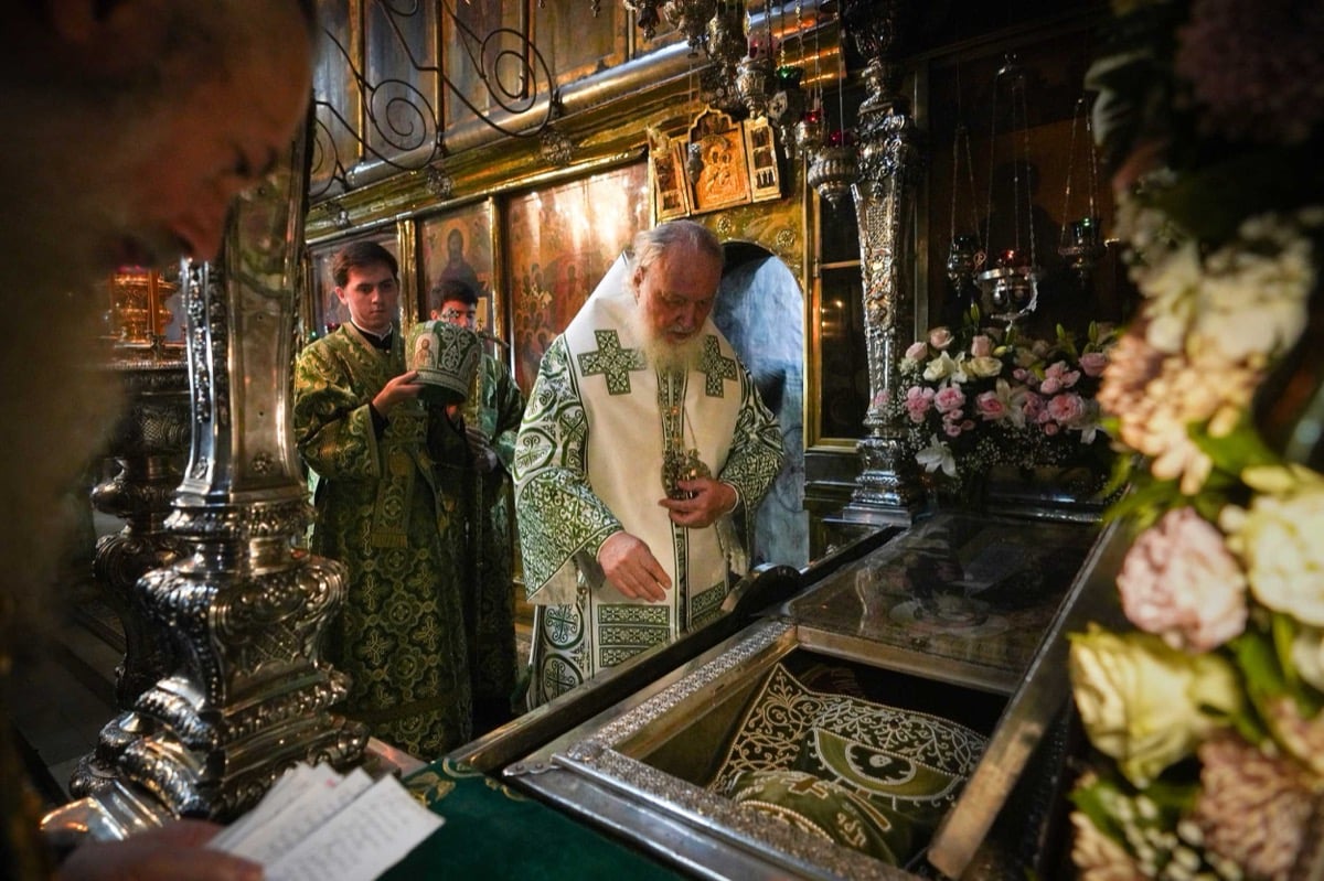 Patriarch Kirill in green vestments venerating the relics of St. Sergius of Radonezh inside the Trinity Cathedral at the Lavra, bowing over the open silver reliquary surrounded by flowers and oil lamps