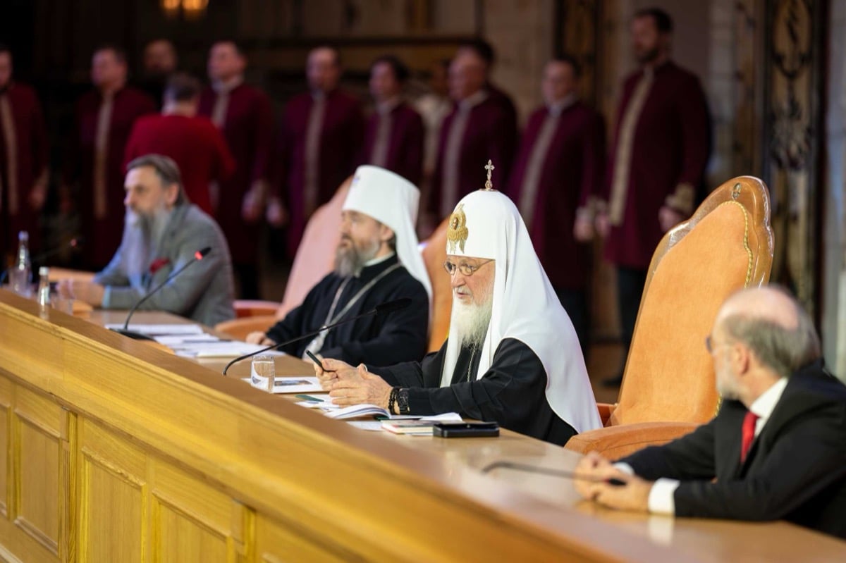 Patriarch Kirill speaking at the WRPC presidium, pen in hand, flanked by a metropolitan and suited officials, with a choir in burgundy vestments behind