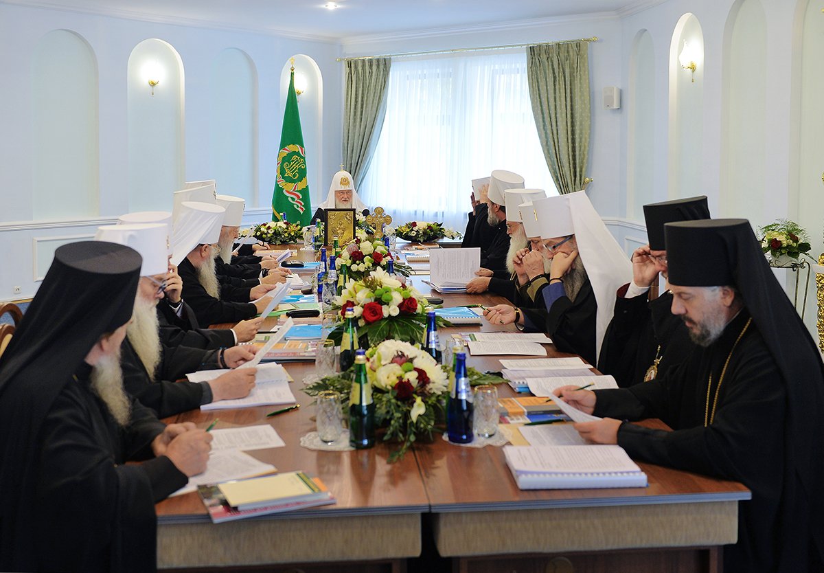 Patriarch Kirill presiding at the Holy Synod of the Russian Orthodox Church in October 2018, with bishops seated around a long conference table holding documents and a central icon