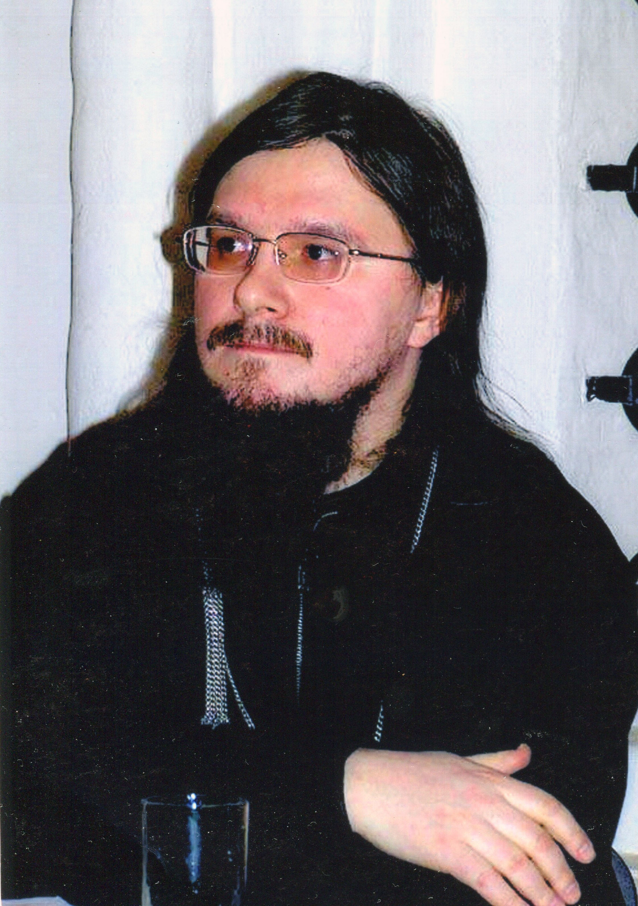 Portrait of Hieromartyr Daniel Sysoev, Russian Orthodox priest with long dark hair, glasses, and a dark cassock, seated with arms crossed
