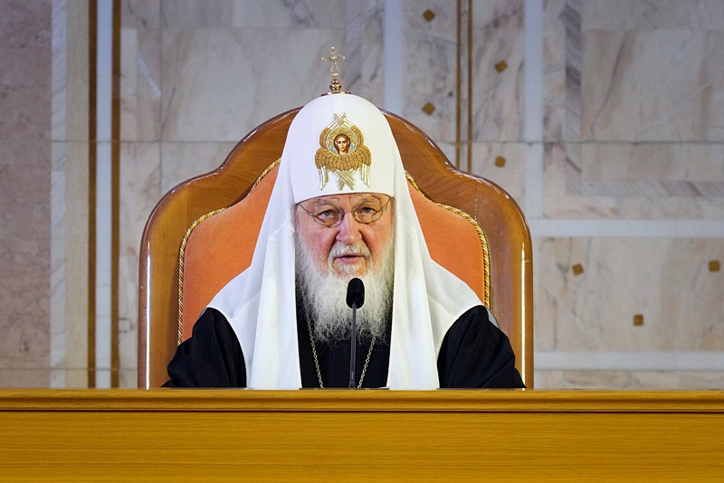 Patriarch Kirill speaking from his patriarchal throne at a formal assembly of Russian Orthodox bishops in a large paneled hall, November 2024