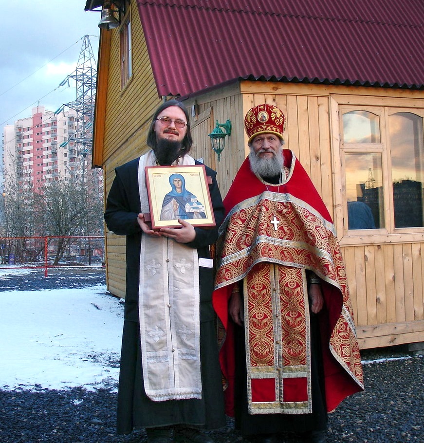 Hieromartyr Daniel Sysoev (left) holding an icon, standing with a fellow priest outside a wooden church in Moscow